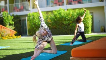 Yoga at The Saguaro Palm Springs, a gay-friendly hotel. Two women on yoga mats on the lawn, exercising. Green grass and colorful building.