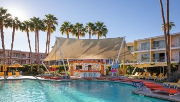 The Saguaro Palm Springs gay-friendly hotel pool view. Palm trees, colorful buildings, bar, and lounge chairs surround the pool. Tropical vibe.