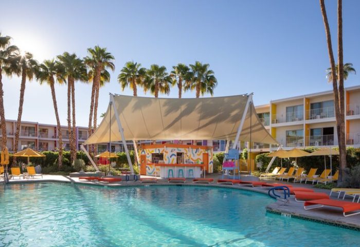 The Saguaro Palm Springs gay-friendly hotel pool view. Palm trees, colorful buildings, bar, and lounge chairs surround the pool. Tropical vibe.