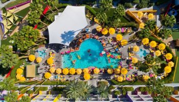 Aerial view of The Saguaro Palm Springs, a gay-friendly hotel. People enjoy the pool, yellow umbrellas, and colorful hotel architecture.