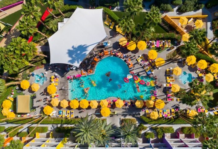 Aerial view of The Saguaro Palm Springs, a gay-friendly hotel. People enjoy the pool, yellow umbrellas, and colorful hotel architecture.