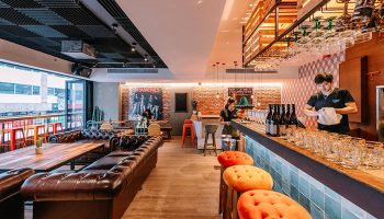 Interior of The Sheung Wan, a gay-friendly hotel in Hong Kong. The bar area features leather seating, a brick wall, and a bartender.