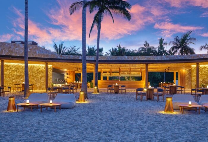 Gay-Friendly Hotel The St. Regis Punta Mita Resort beach bar at dusk. Tables and chairs on the sand, palm trees, and a colorful sky.