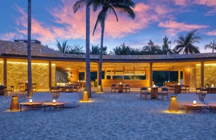 Gay-Friendly Hotel The St. Regis Punta Mita Resort beach bar at dusk. Tables and chairs on the sand, palm trees, and a colorful sky.
