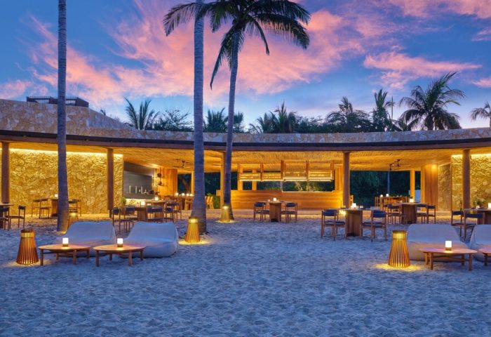 Gay-Friendly Hotel The St. Regis Punta Mita Resort beach bar at dusk. Tables and chairs on the sand, palm trees, and a colorful sky.