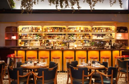 Interior of The Standard East Village, a gay-friendly hotel. Bar with stools, tables, and liquor bottles. Plants hang from the ceiling.