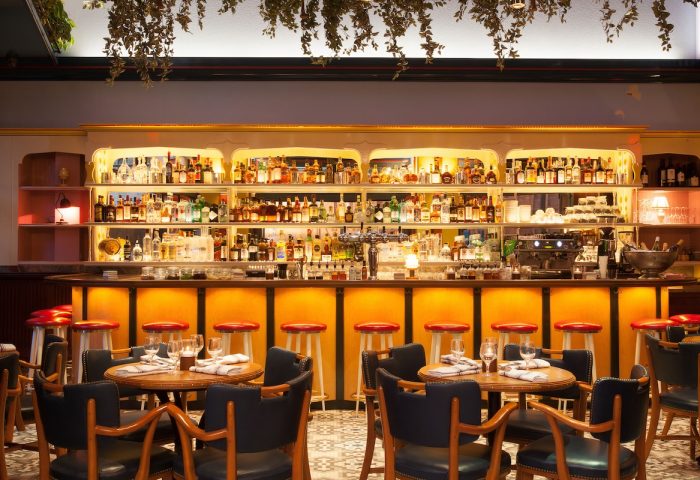 Interior of The Standard East Village, a gay-friendly hotel. Bar with stools, tables, and liquor bottles. Plants hang from the ceiling.