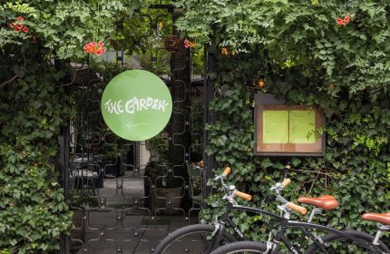 The Standard East Village hotel's entrance, a gay-friendly space. Bicycles parked outside The Garden sign, lush greenery surrounds the gate.