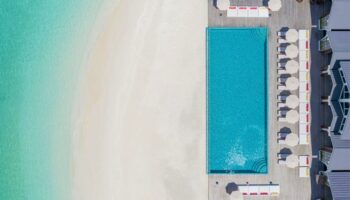 Aerial view of The Standard, Huruvalhi Maldives, a gay-friendly hotel. Pool, beach, umbrellas, and lounge chairs visible. Tropical paradise.