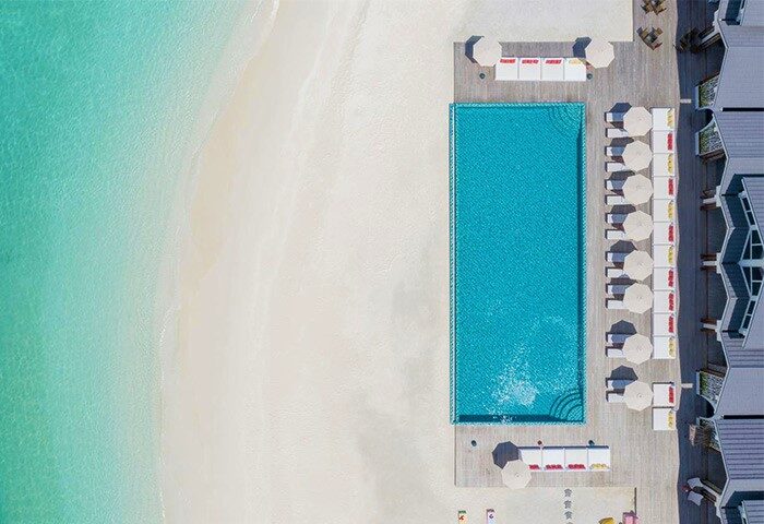 Aerial view of The Standard, Huruvalhi Maldives, a gay-friendly hotel. Pool, beach, umbrellas, and lounge chairs visible. Tropical paradise.