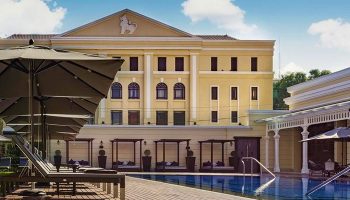 The Strand Yangon gay-friendly hotel pool. Loungers, umbrellas, and cabanas line the pool's edge. Yellow building in the background.