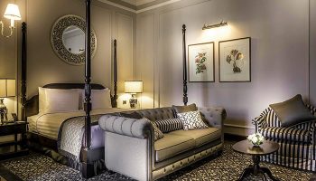 Gay-Friendly Hotel The Strand Yangon: Elegant room with four-poster bed, sofa, and armchair. A luxurious stay in Yangon, Myanmar.