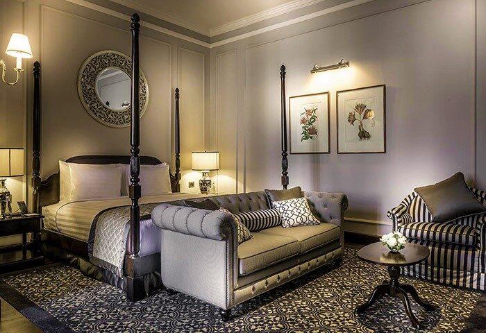 Gay-Friendly Hotel The Strand Yangon: Elegant room with four-poster bed, sofa, and armchair. A luxurious stay in Yangon, Myanmar.