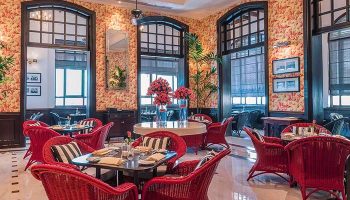 The Strand Yangon, a gay-friendly hotel. Interior view of the restaurant with red wicker chairs, floral wallpaper, and large windows.