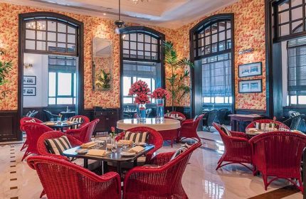 The Strand Yangon, a gay-friendly hotel. Interior view of the restaurant with red wicker chairs, floral wallpaper, and large windows.