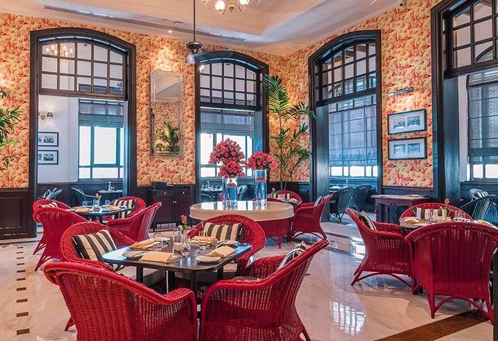 The Strand Yangon, a gay-friendly hotel. Interior view of the restaurant with red wicker chairs, floral wallpaper, and large windows.