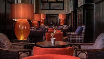 Interior of The Strand Yangon, a gay-friendly hotel. Lamps with orange shades, plaid furniture, and dark wood create a warm, luxurious ambiance.