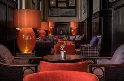 Interior of The Strand Yangon, a gay-friendly hotel. Lamps with orange shades, plaid furniture, and dark wood create a warm, luxurious ambiance.