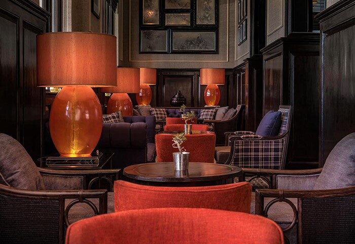 Interior of The Strand Yangon, a gay-friendly hotel. Lamps with orange shades, plaid furniture, and dark wood create a warm, luxurious ambiance.