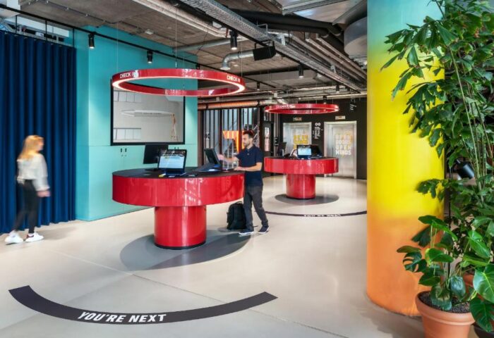 The Student Hotel Amsterdam City lobby. Modern, gay-friendly hotel check-in area with red desks and You're Next floor markings. Colorful decor.