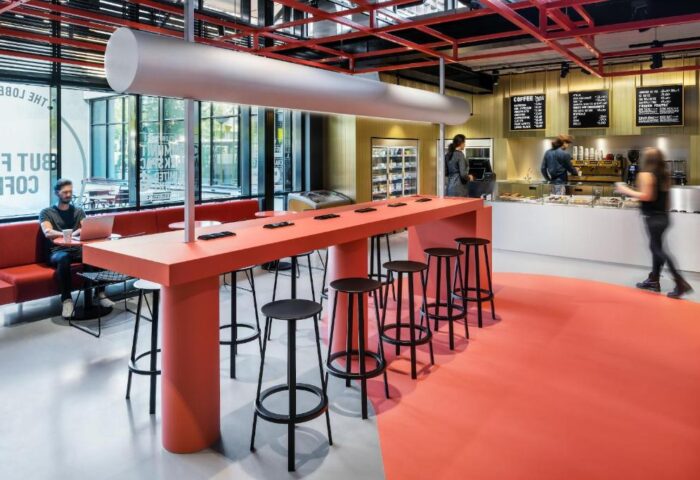 The Student Hotel Amsterdam City interior. Gay-friendly hotel's shared workspace with red table, stools, and cafe. Modern design, vibrant colors.