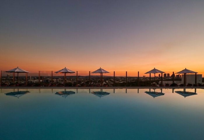 Sunset at The West Hollywood EDITION, a gay-friendly hotel. Umbrellas line the rooftop pool, reflecting in the water. Skyline view.