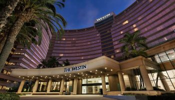 The Westin Long Beach hotel exterior at dusk. Gay-friendly hotel with palm trees. Entrance to The Westin is lit up.