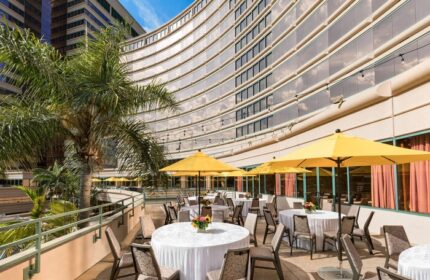 Outdoor patio at The Westin Long Beach, a gay-friendly hotel. Tables with white linens and yellow umbrellas. Palm tree and modern architecture.