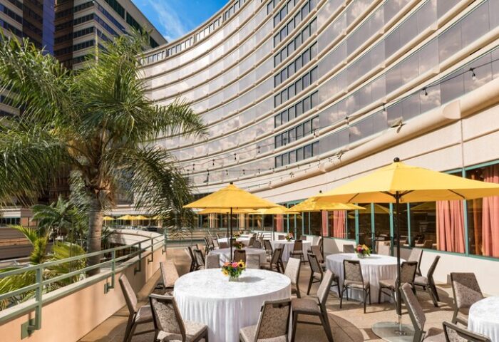 Outdoor patio at The Westin Long Beach, a gay-friendly hotel. Tables with white linens and yellow umbrellas. Palm tree and modern architecture.