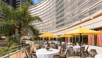 Outdoor patio at The Westin Long Beach, a gay-friendly hotel. Tables with white linens and yellow umbrellas. Palm tree and modern architecture.