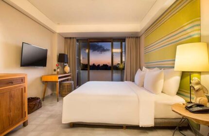 Tijili Hotel Seminyak, gay-friendly hotel. Room interior with a large bed, TV, desk, and balcony view at sunset. Modern design.