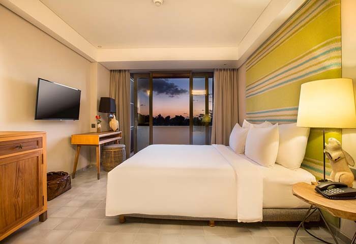Tijili Hotel Seminyak, gay-friendly hotel. Room interior with a large bed, TV, desk, and balcony view at sunset. Modern design.