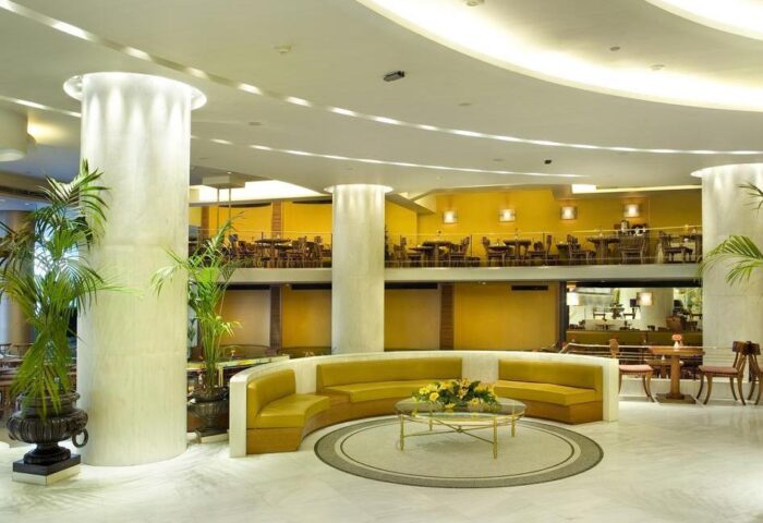 Titania Hotel's lobby: Gay-friendly hotel with a modern design, featuring a seating area, pillars, plants, and a restaurant on the upper floor.