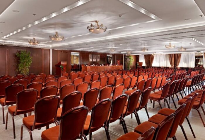 Conference room at Titania Hotel, a gay-friendly hotel. Rows of red chairs face a stage. Ideal for events and gatherings.
