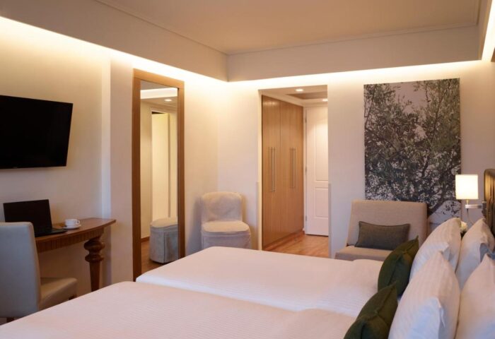 Titania Hotel gay-friendly room. Two beds with white linens, desk with laptop, TV, closet, mirror, and olive tree art. Modern, clean design.