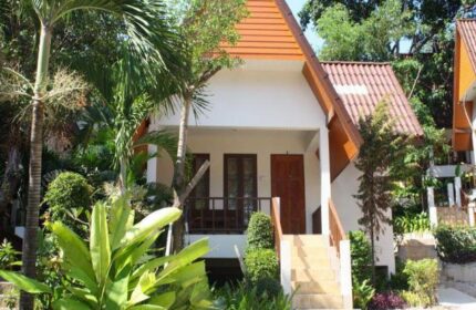 Tok Little Hut, a gay-friendly hotel. Exterior view of a charming bungalow with a peaked roof, surrounded by lush tropical greenery. Welcoming stairs.