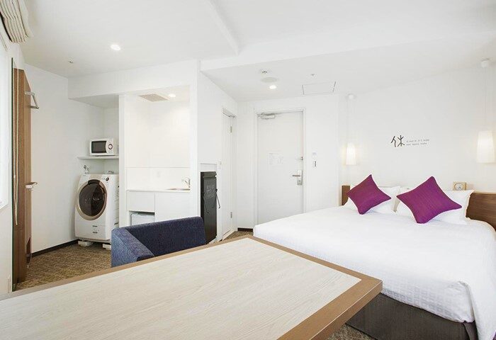 Tokyu Stay Shinjuku, gay-friendly hotel room. Includes bed with purple pillows, table, microwave, and washing machine. Modern, clean design.