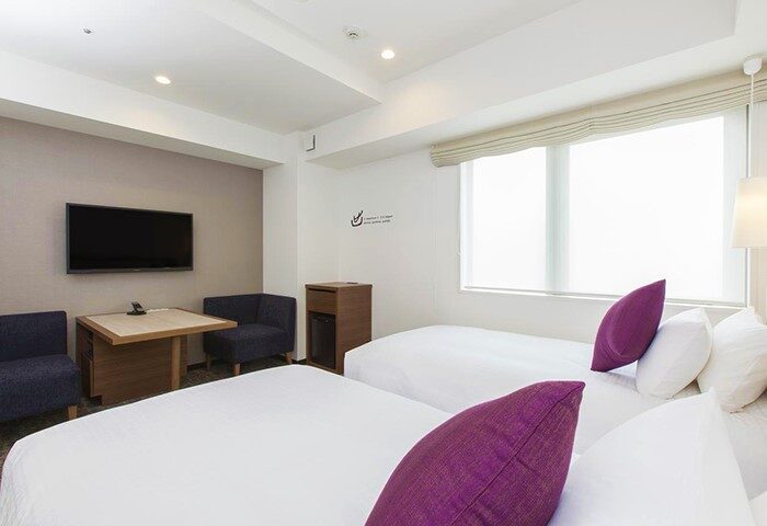 Tokyu Stay Shinjuku room. Gay-friendly hotel with two beds, purple pillows, TV, table, and chairs. Modern design for gay travelers in Shinjuku.