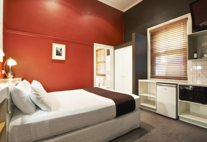Tolarno Hotel room. Gay-friendly hotel with a bed, red wall, wardrobe, window with blinds, microwave, and mini-fridge. Gay travel advice.