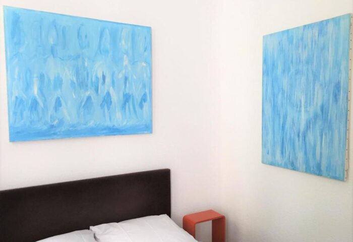 Tom's Hotel, a gay-friendly hotel: Bedroom with blue abstract paintings, bed with white linens, and a small orange side table. Gay hotel room.