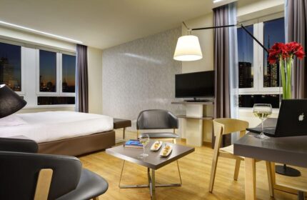 Gay-Friendly UNAHOTELS Century Milano room. Modern interior with bed, chairs, desk, laptop, wine, snacks, and city view. Gay travel.