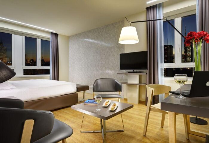 Gay-Friendly UNAHOTELS Century Milano room. Modern interior with bed, chairs, desk, laptop, wine, snacks, and city view. Gay travel.