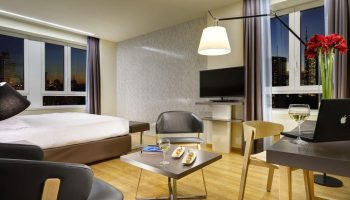 Gay-Friendly UNAHOTELS Century Milano room. Modern interior with bed, chairs, desk, laptop, wine, snacks, and city view. Gay travel.
