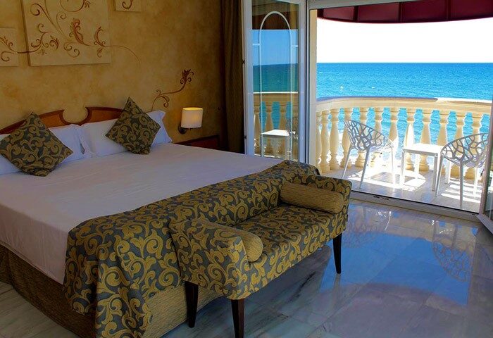 URH Sitges Playa gay-friendly hotel room with balcony ocean view. Pet-friendly room with bed, sofa, and balcony with chairs.