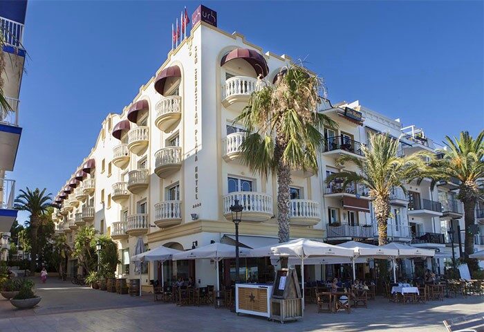 URH Sitges Playa, a gay-friendly, pet-friendly hotel. Exterior view of the hotel with balconies, outdoor dining, and palm trees.