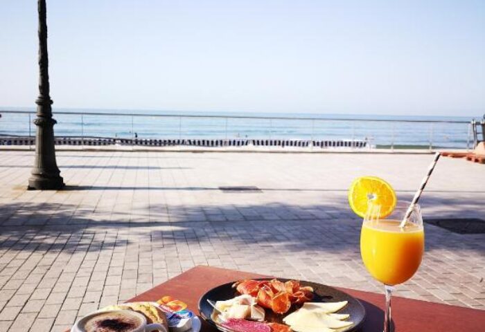Breakfast with sea view at URH Sitges Playa, a gay-friendly, pet-friendly hotel. Croissants, coffee, juice, and cold cuts on a sunny day.