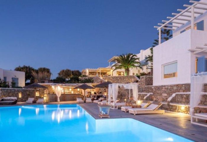 Vencia Boutique Hotel's pool at dusk. A gay-friendly hotel with a bar, lounge chairs, and white buildings on a hill in Mykonos, Greece.