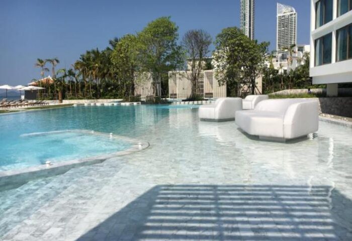 Veranda Resort Pattaya MGallery pool view. Gay-friendly hotel with stylish white lounge seating in the water. Tropical trees, blue sky.