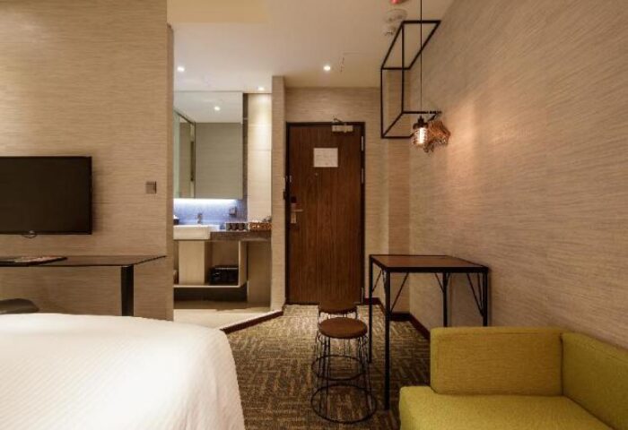 Via Hotel Taipei Station room interior. Gay-friendly hotel with bed, desk, stool, and sofa. Bathroom visible. Modern design.