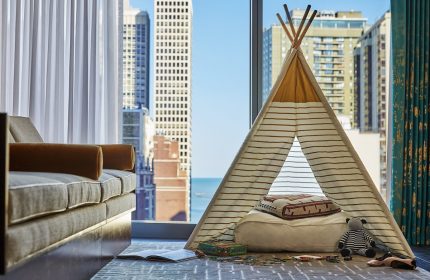 Viceroy Chicago gay-friendly hotel room. A child's teepee tent sits by a window with a city view. Sofa, toys, and a book are nearby.